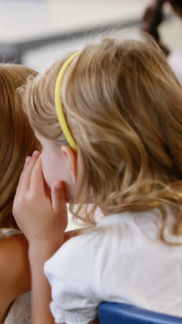 Schoolgirl-whispering-into-her-friend-s-ear-in-classroom