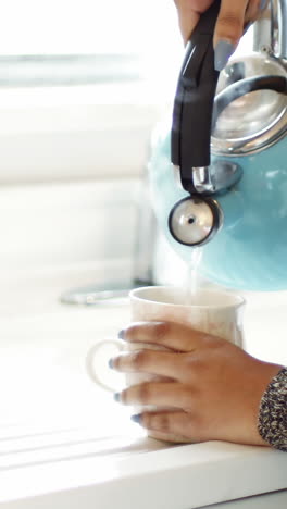 Woman-pouring-hot-water-from-kettle-into-mug-in-kitchen