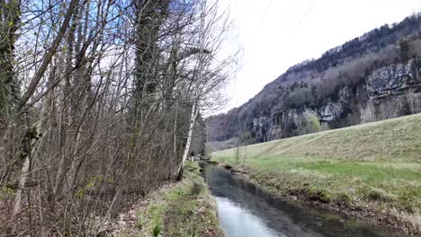 A-white-dog-races-alongside-the-waterway,-set-against-the-grandeur-of-Switzerland's-majestic-mountain-ranges,-symbolizing-the-essence-of-freedom-and-the-allure-of-natural-beauty