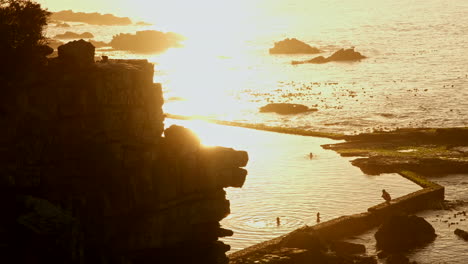 Bright-golden-hour-sunrise-light-over-ocean-and-marine-tide-pool-on-coastline-in-Hermanus-with-people-swimming