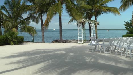 Wide-panning-shot-from-tree-to-wedding-venue-decor-and-flowers-outdoor-seating-sandy-beach-engagement-seating-with-tables