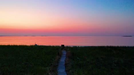 Wooden-Walking-Path-Leading-to-Colorful-Ocean-Sunset-on-Beach-with-Trees-and-Grass-at-Dusk-with-Beautiful-Red,-Orange,-and-Purple-Colors-in-the-Sky-and-Reflecting-off-Water