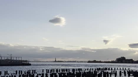 Ein-Ruhiger-Blick-Auf-Liberty-Island-Bei-Sonnenuntergang,-Eingerahmt-Vom-Ruhigen-Wasser-Und-Den-Verstreuten-Pfählen-Des-Hafens,-Mit-Der-Freiheitsstatue-Als-Silhouette-Gegen-Den-Himmel