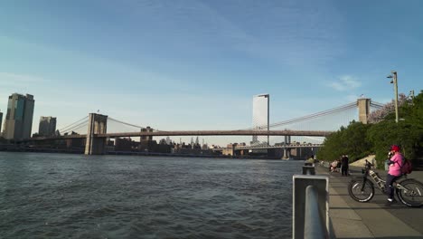 Blick-Auf-Den-East-River-Mit-Brooklyn-Bridge-Und-Manhattan-Bridge-Im-Hintergrund-Mit-Menschen-Auf-Dem-Wanderweg