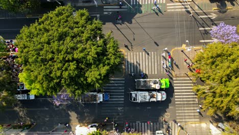 Traveling-left-overhead-aerial-view-of-a-group-of-garbage-trucks-accompanying-a-pilgrimage-in-Mexico-City-on-a-sunny-day,-wooded-area