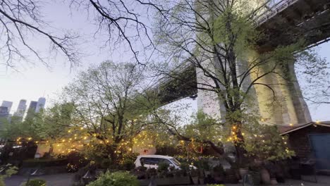 A-unique-perspective-from-beneath-the-Brooklyn-Bridge,-looking-up-through-the-trees-with-string-lights,-capturing-the-bridge's-structure-against-a-clear-evening-sky