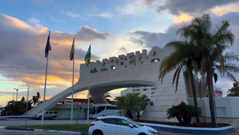 Big-Marbella-sign-with-flags-and-sunset-moving-orange-clouds-in-Spain,-beautiful-sky-and-palm-trees,-cars-driving-on-a-road,-4K-static-shot