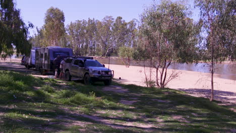 A-young-family-sets-up-camp-next-to-a-river-with-sandy-banks,-enjoying-a-peaceful-getaway-in-their-caravan