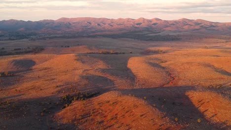 Spektakuläre-Drohnenaufnahme-Eines-Tiefflugs-über-Den-Sanften-Hügeln-Des-Australischen-Outbacks,-Während-Die-Sonne-über-Den-Flinders-Ranges-Untergeht