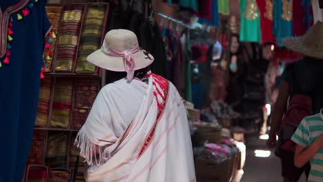 An-old-lady-wearing-a-traditional-outfit-and-a-hat-is-shopping-for-clothing-in-a-store-with-many-other-people-around-her