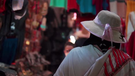 An-old-lady-wearing-a-traditional-outfit-and-a-straw-hat-with-pink-ribbon-around-it-is-standing-in-front-of-a-store-filled-with-clothing-and-accessories