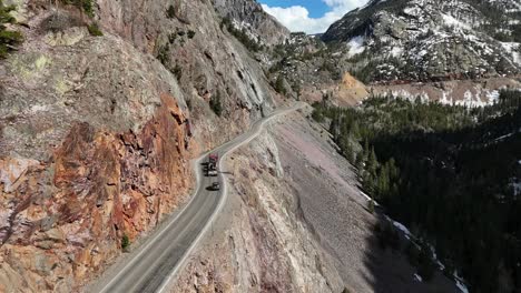 Semi-Truck-Driving-on-the-edge-of-a-cliff-with-a-huge-drop-off,-surrounded-by-mountains,-pine-trees-at-bottom-of-cliff,-spring-in-Ouray-Colorado,-red-mountains-pass,-Million-Dollar-Highway