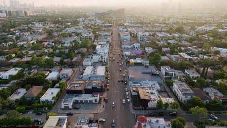 Drone-Shot-Flying-over-Melrose-Avenue-in-Los-Angeles,-California-in-Daytime-with-Cars-Driving-Below-and-DTLA-Skyline-on-Horizon