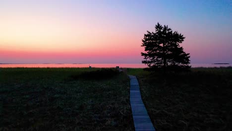 POV-Walk-on-Wooden-Walking-Path-Leading-to-Colorful-Ocean-Sunrise-on-Beach-with-Trees-and-Grass-at-Dawn-with-Beautiful-Red,-Orange,-and-Purple-Colors-in-the-Sky-and-Reflecting-off-Water