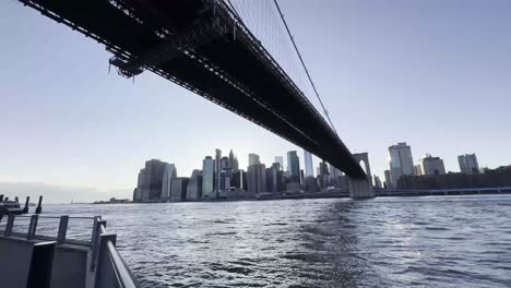 Ein-Atemberaubender-Blick-In-Der-Dämmerung-Unter-Der-Brooklyn-Bridge,-Der-Die-Weite-Des-East-River-Und-Die-Beleuchtete-Skyline-Von-Manhattan-Einfängt-Und-Die-Lebendige-Abendatmosphäre-Der-Stadt-Widerspiegelt