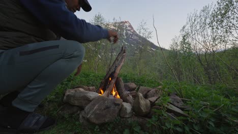 El-Hombre-Pone-Un-Palo-Extra-En-La-Hoguera-Y-Sostiene-Las-Manos-Encima-Para-Calentarse,-Vista-A-La-Montaña