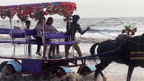 Los-Hombres-Montan-A-Caballo-En-La-Playa,-Los-Carruajes-Tirados-Por-Caballos-Van-Bajo-El-Agua.