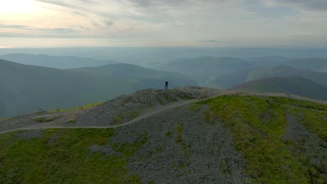 Solitary-mountain-walker-on-summit-with-wide-slow-orbit-revealing-misty-valleys-and-mountains-prior-to-sunset