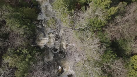 A-rocky-waterfall-surrounded-by-lush-and-bare-trees-in-owen-sound,-canada,-aerial-view