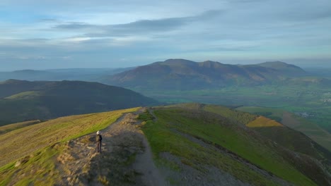 Flying-over-golden-hour-lit-mountain-summit-with-lone-walker-and-towards-distant-Skiddaw-mountain-range