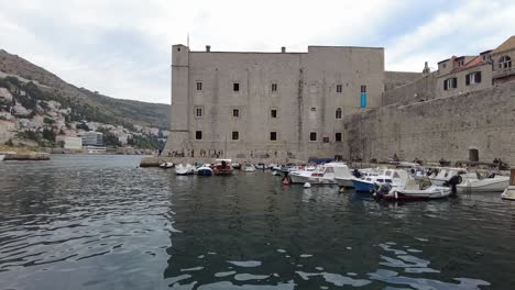 Panoramic-daytime-view-of-Dubrovnik,-Croatia,-showcasing-boats-docked-at-the-small-harbor-and-people-passing-by-the-historic-fortress-wall