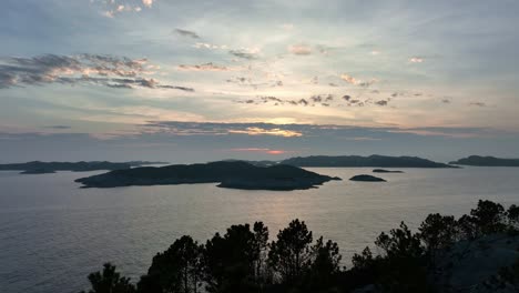 Drone-moves-over-silhouetted-pine-treetops-towards-the-North-Sea,-coastal-islands,-and-a-stunning-sunset-sky