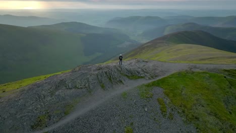 Lone-walker-on-mountain-summit-staring-at-misting-fells-and-valleys-beyond-during-golden-hour