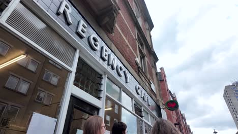 Low-Angle-Looking-Up-At-Iconic-Regency-Cafe-Sign-With-People-Queuing-Outside-Waiting-To-Get-A-Table