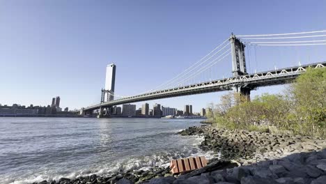 Manhattan-Bridge-spanning-across-the-East-River,-with-the-rocky-riverbank-and-the-New-York-City-skyline-in-the-background,-capturing-the-tranquility-and-beauty-of-the-city's-waterfront