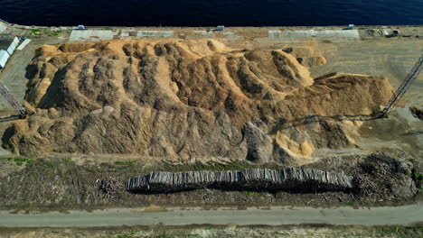 Tree-trunks-and-large-pile-of-wood-fibers-heaped-up-at-the-harbor,-aerial-slider