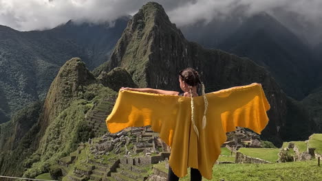 Woman-raises-arms-wearing-yellow-poncho-in-front-of-Machu-picchu,-rearview