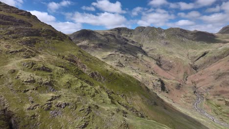 Rocky-green-mountainside-with-sunlit-Crinkle-Crag-mountain-range-beyond-under-bright-blue-cloudy-sky-and-pan-showing-Bow-Fell