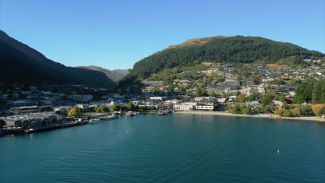 Low,-wide-aerial-flying-towards-beach,-buildings-of-Queenstown,-New-Zealand