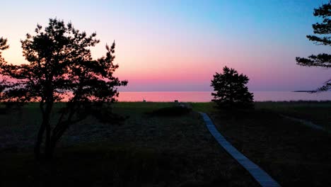 Holzweg,-Der-Zu-Einem-Farbenfrohen-Sonnenaufgang-Am-Strand-Mit-Bäumen-Und-Gras-Im-Morgengrauen-Führt,-Mit-Wunderschönen-Roten,-Orangen-Und-Violetten-Farben-Am-Himmel-Und-Spiegelungen-Im-Wasser