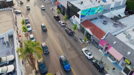 Toma-Aérea-De-Automóviles-Conduciendo-Por-La-Avenida-Melrose-Durante-El-Día,-Mirando-Hacia-Abajo-A-Los-Peatones-Caminando-Por-La-Acera-Junto-A-Las-Vibrantes-Tiendas-De-West-Hollywood