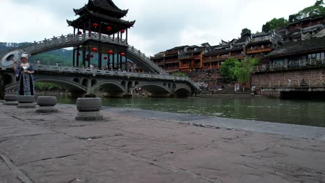 Low-angle-shot-of-Miao-ethnic-girl-with-umbrella-in-front-of-Snow-bridge-in-Fenghuang,-China