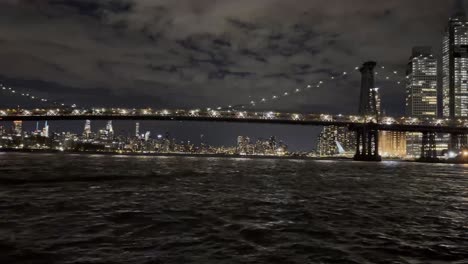 Ein-Friedlicher-Nächtlicher-Blick-Auf-Den-East-River-Mit-Der-Beleuchteten-Manhattan-Bridge,-Die-Sich-Im-Wasser-Spiegelt,-Und-Der-Skyline-Von-New-York-Im-Hintergrund-Unter-Einem-Bewölkten-Himmel