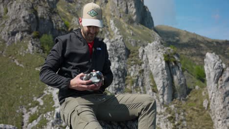 Hombre-Sacando-Un-Dron-Doblado-Del-Bolsillo-De-La-Chaqueta-Y-Desplegando-Los-Brazos-Preparándolo-Para-Volar-Con-Fondo-De-Montaña