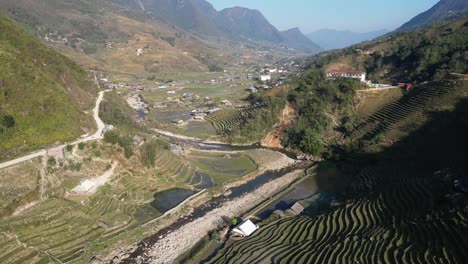 Aerial-drone-shot-of-road-leading-through-villages-amidst-bright-green-rice-terraces-in-the-mountains-of-Sapa,-Vietnam