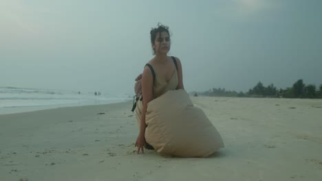 A-woman-in-a-light-dress,-with-her-hair-in-a-bun,-kneels-on-a-serene-beach-at-dusk,-drawing-in-the-sand-with-her-finger