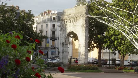 Los-Coches-Circulan-Por-La-Calle-Cerca-De-Un-Arco-Triunfal,-La-Antigua-Arquitectura-Europea-En-Un-Parque-Con-árboles-Y-Flores