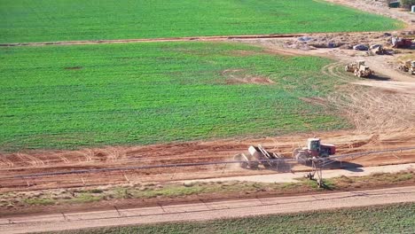 Tractor-and-small-road-grader-driving-on-farm-dirt-road-beside-other-farm-equipment