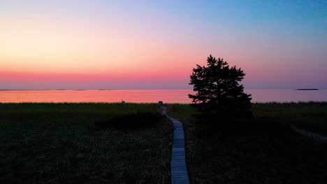 Caminata-Pov-Por-Un-Sendero-De-Madera-Que-Conduce-A-Una-Colorida-Puesta-De-Sol-En-El-Océano-En-Una-Playa-Con-árboles-Y-Pasto-Al-Atardecer-Con-Hermosos-Colores-Rojo,-Naranja-Y-Morado-En-El-Cielo-Y-Reflejándose-En-El-Agua