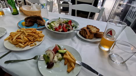 Variedad-De-Platos-Griegos-Que-Incluyen-Ensalada,-Papas-Fritas-Y-Cerveza-En-La-Mesa-Del-Restaurante-Al-Aire-Libre.