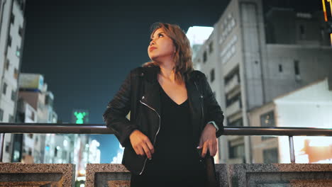 Asian-woman-in-black-leather-jacket-stands-on-a-bridge-at-night,-gazing-at-the-vibrant-city-lights