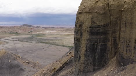 Aerial-view-of-a-towering-butte-in-Moab,-Utah,-with-expansive-desert-and-canyon-landscape-in-the-background,-showcasing-the-rugged-and-dramatic-terrain-under-a-cloudy-sky