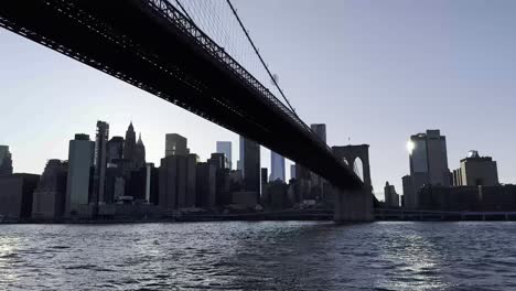 A-captivating-view-from-beneath-the-Brooklyn-Bridge-at-dusk,-highlighting-the-silhouette-of-Manhattan's-skyline-against-the-twilight-sky-and-the-shimmering-waters-of-the-East-River