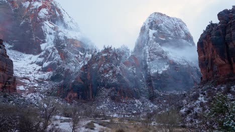 Pan-up-shot-showcasing-a-snow-covered-field-with-trees-and-shrubs-growing-below-the-towering-red-rock-mountains-in-Zion-National-Park