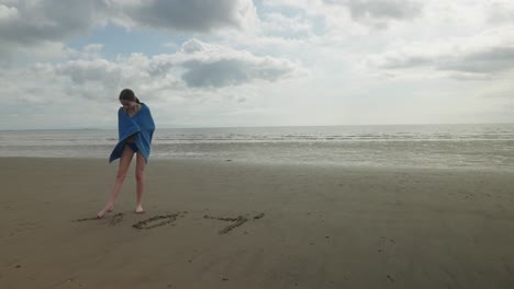 Teenage-girl-towel-wrapped,-creative-writing,-sandy-beach,-slow-motion