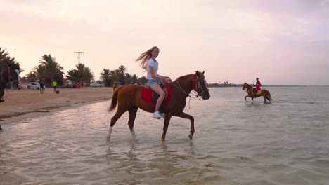 A-woman-rides-a-horse-through-shallow-water-near-a-beach-in-Tunisia,-Djerba